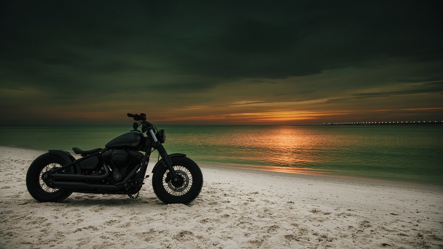 A matte black motorcycle parked on white sand, facing an emerald gulf at sunset, pier lights on the horizon