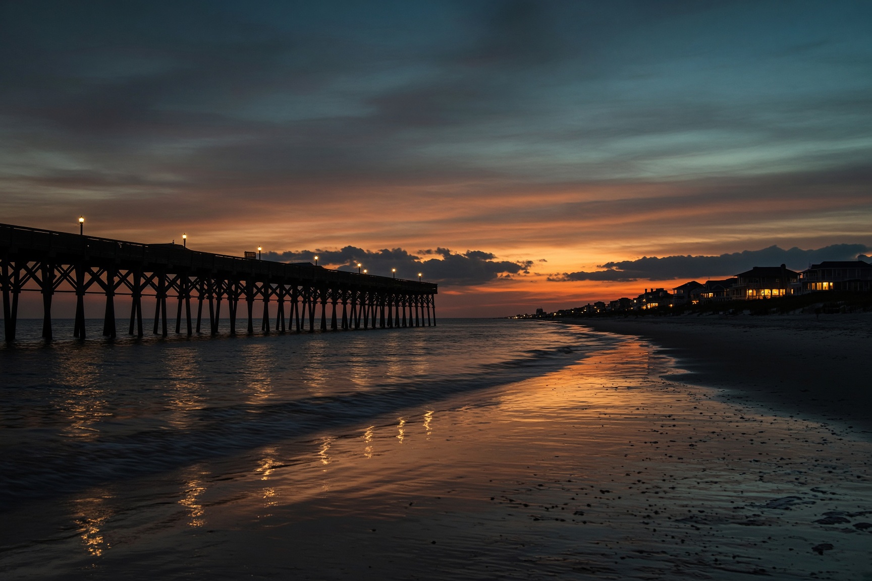 The pier stretching into a copper and teal dusk, lights mirrored on wet sand